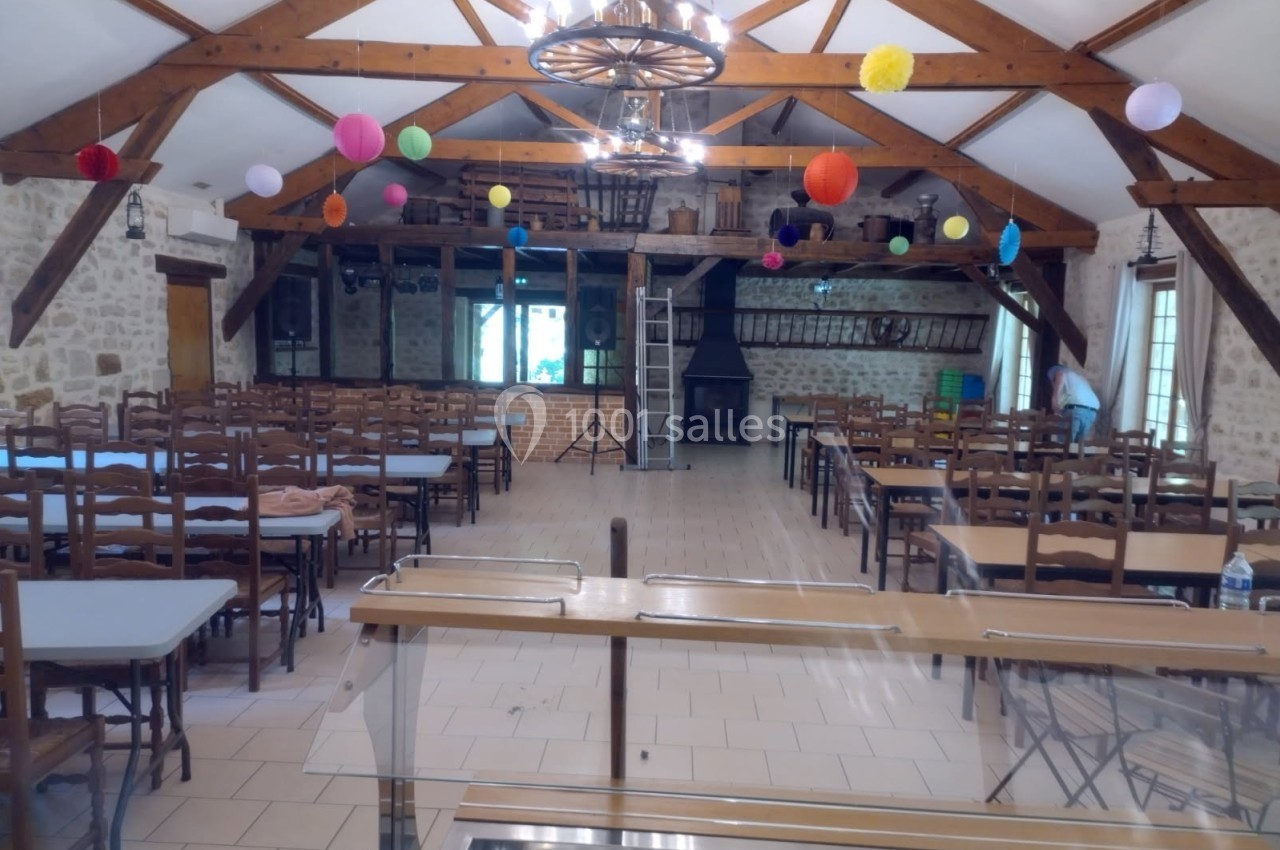 Salle de réception avec poutres apparentes, tables et chaises alignées, décorée de boules colorées suspendues.
