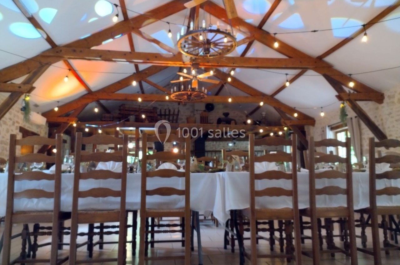 Salle de réception avec poutres apparentes, tables dressées et chaises en bois sous des guirlandes lumineuses.