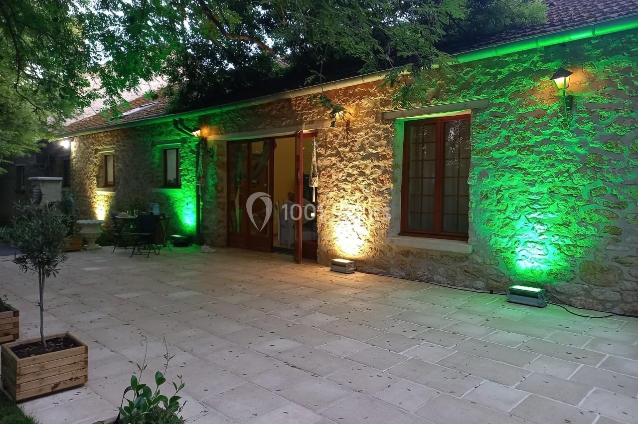 Façade en pierre d'une maison éclairée par des lumières vertes et blanches, avec une terrasse pavée et des plantes en pots.