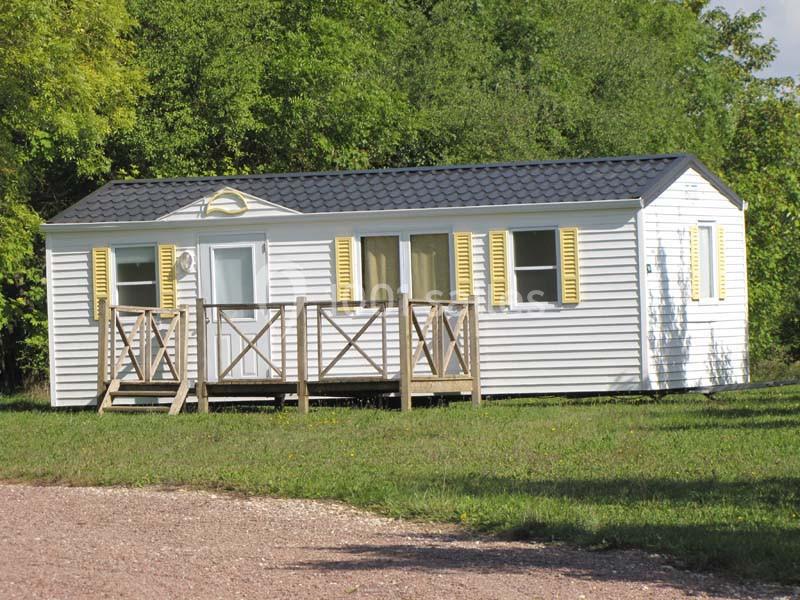 Mobil-home blanc avec volets jaunes, terrasse en bois et toit sombre, situé sur une pelouse entourée d'arbres.
