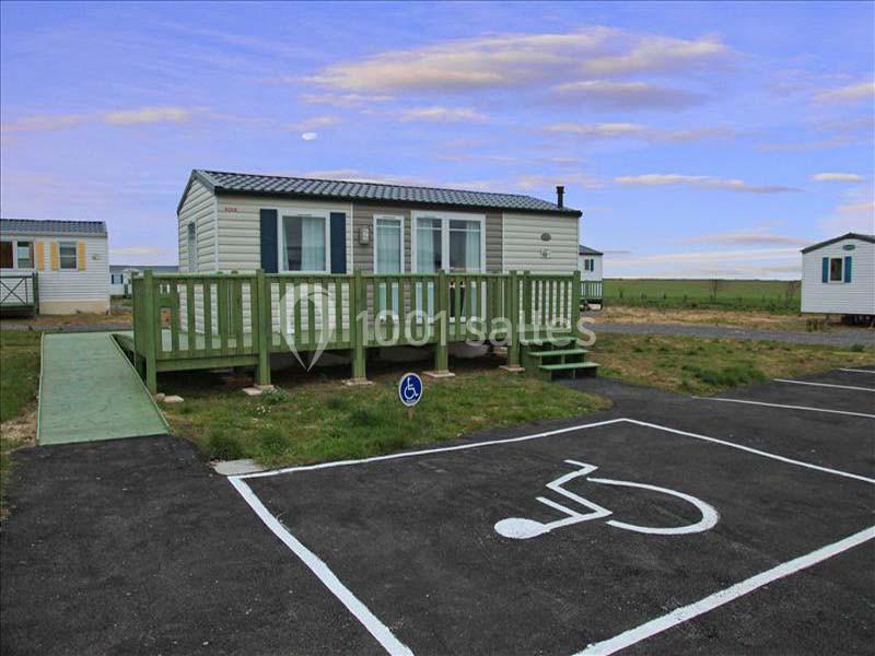 Mobil-home avec rampe d'accès, terrasse en bois et place de parking réservée aux personnes à mobilité réduite.