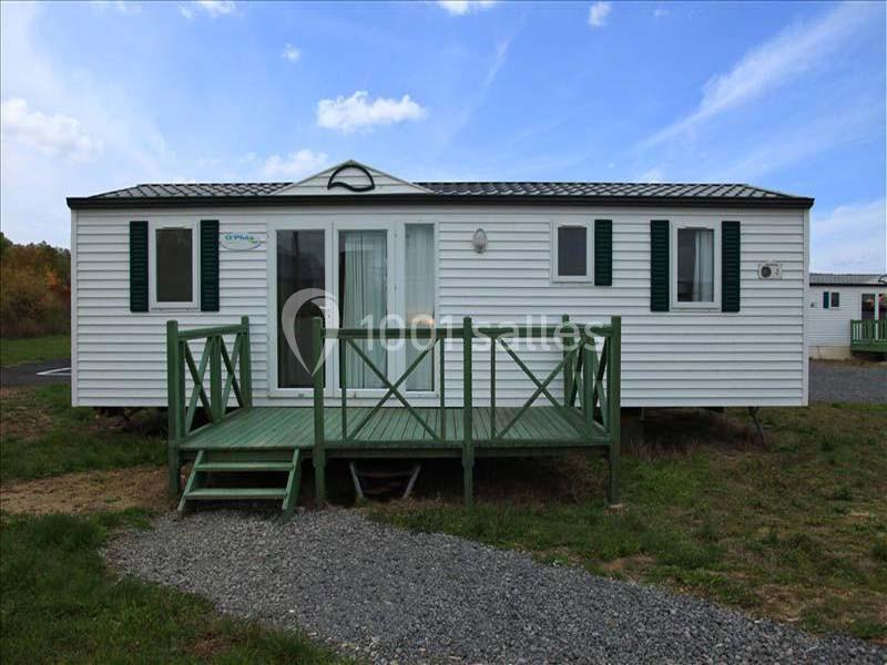 Mobil-home blanc avec terrasse en bois vert, situé sur un terrain herbeux sous un ciel partiellement nuageux.