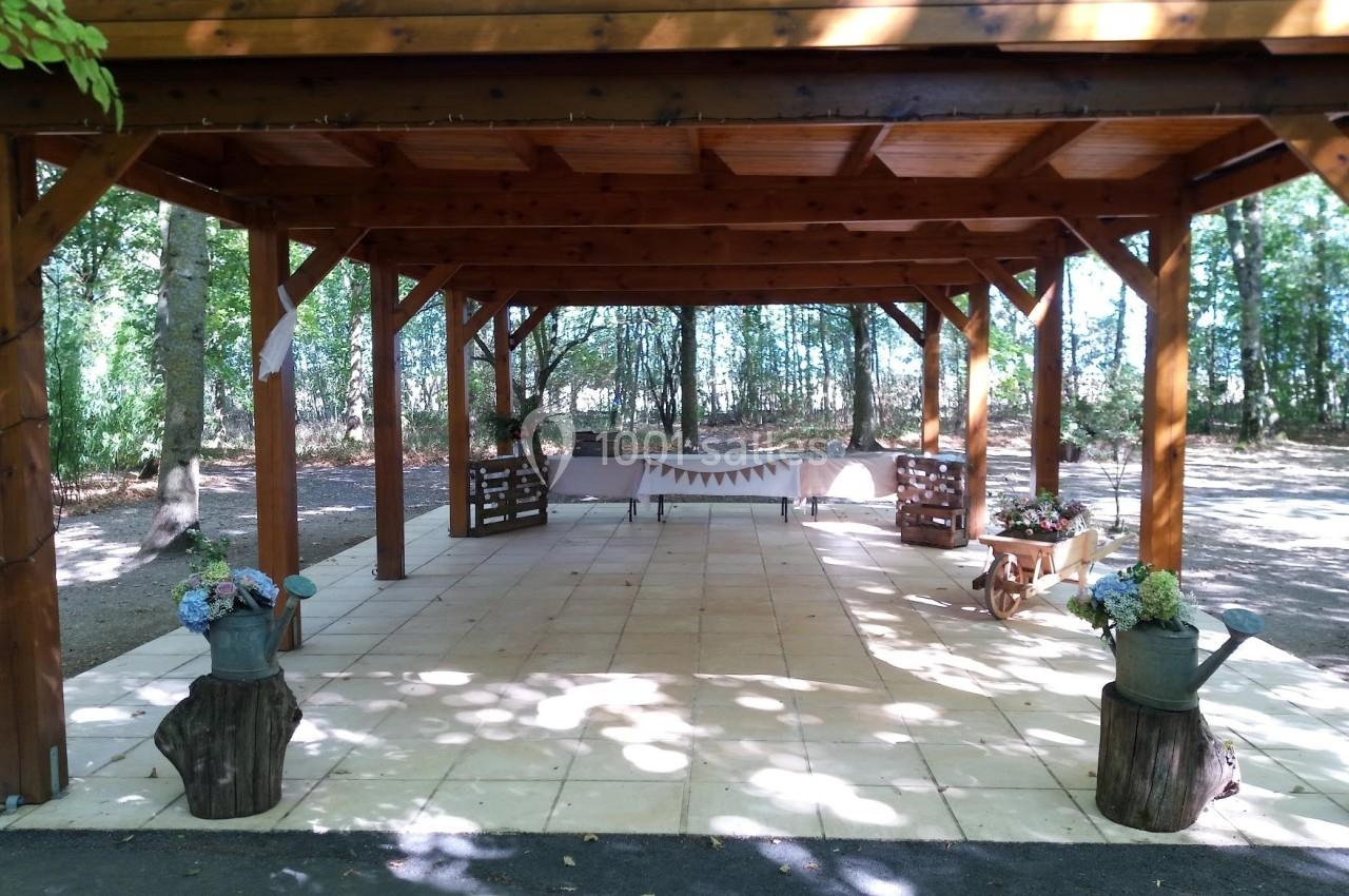 Structure en bois ouverte sur une terrasse carrelée, décorée de fleurs et d'éléments champêtres, entourée d'arbres.