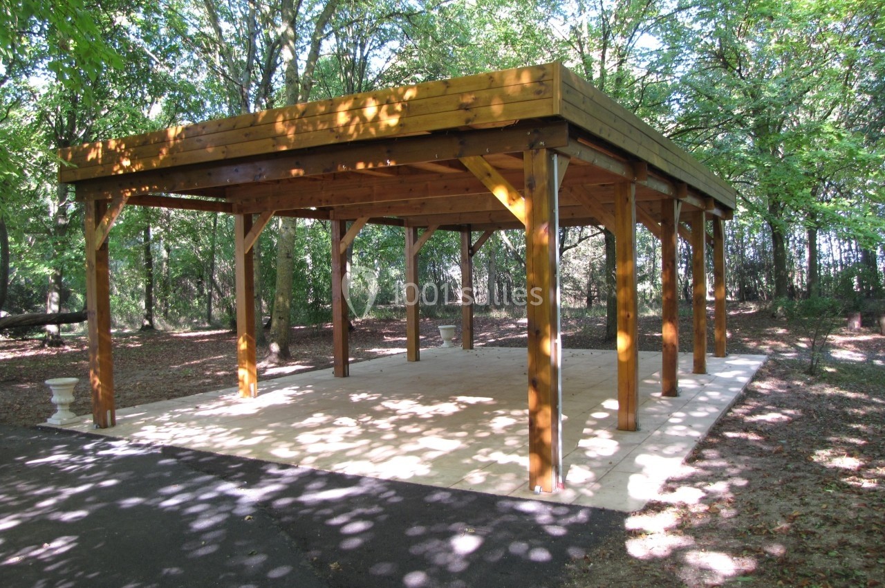 Structure en bois ouverte avec toit plat, située dans une clairière entourée d'arbres.