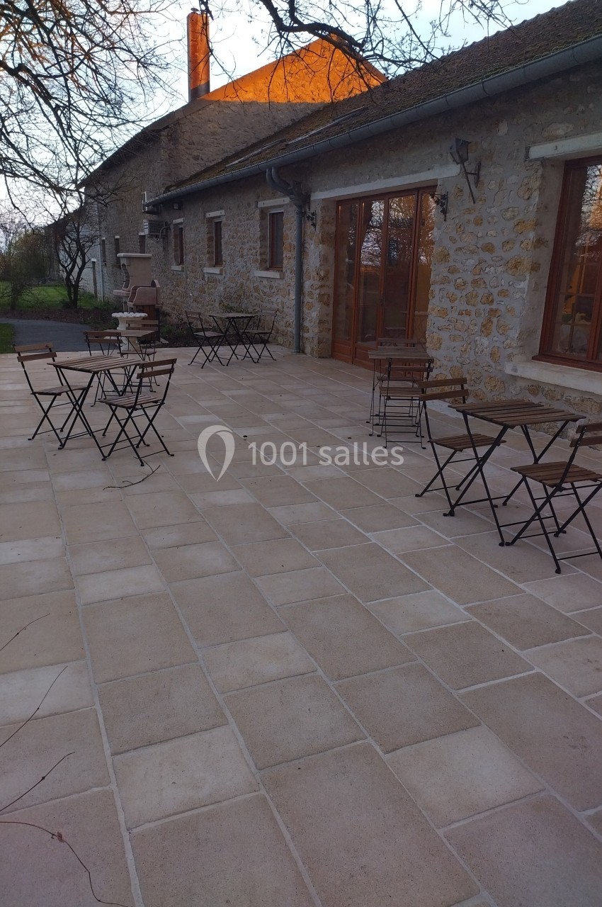 Terrasse en pierre avec tables et chaises en métal devant une maison en pierre, entourée d'arbres au coucher du soleil.