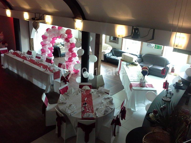 Salle décorée pour un événement avec des tables dressées, des nappes blanches, des rubans rouges et une arche de ballons…