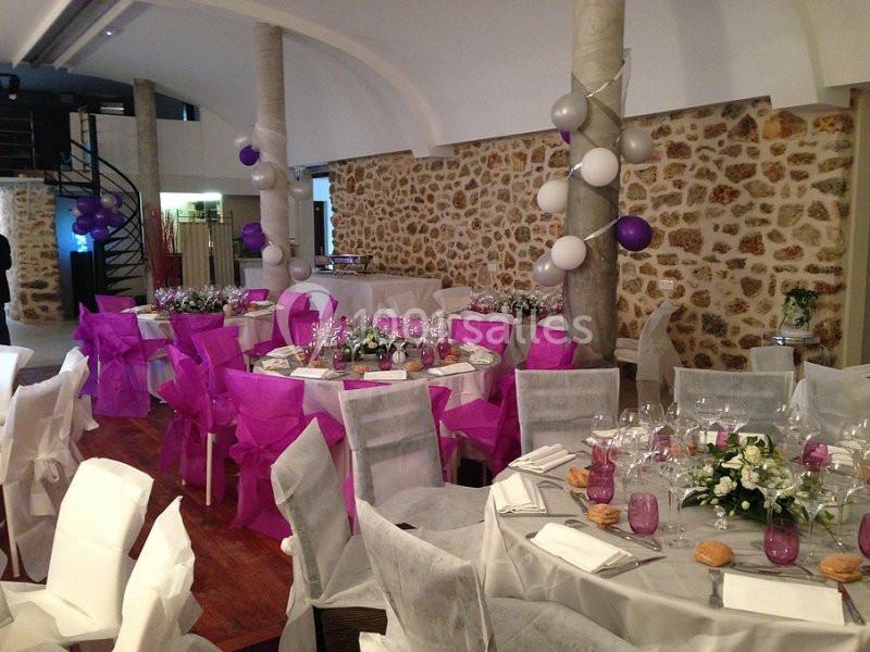 Salle de réception décorée pour un événement, avec tables dressées, chaises ornées de nœuds roses et ballons suspendus.