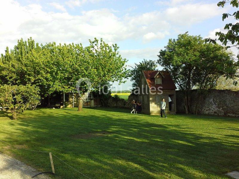 Jardin verdoyant avec pelouse, arbres, petite cabane et deux personnes se tenant près d'un mur en pierre.