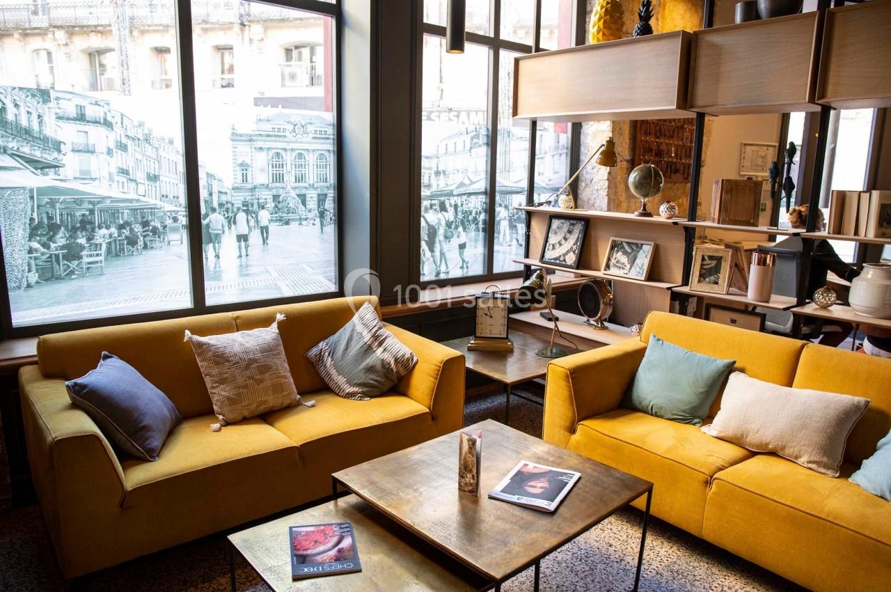 Deux canapés jaunes avec coussins entourent une table basse dans un salon lumineux avec de grandes fenêtres.