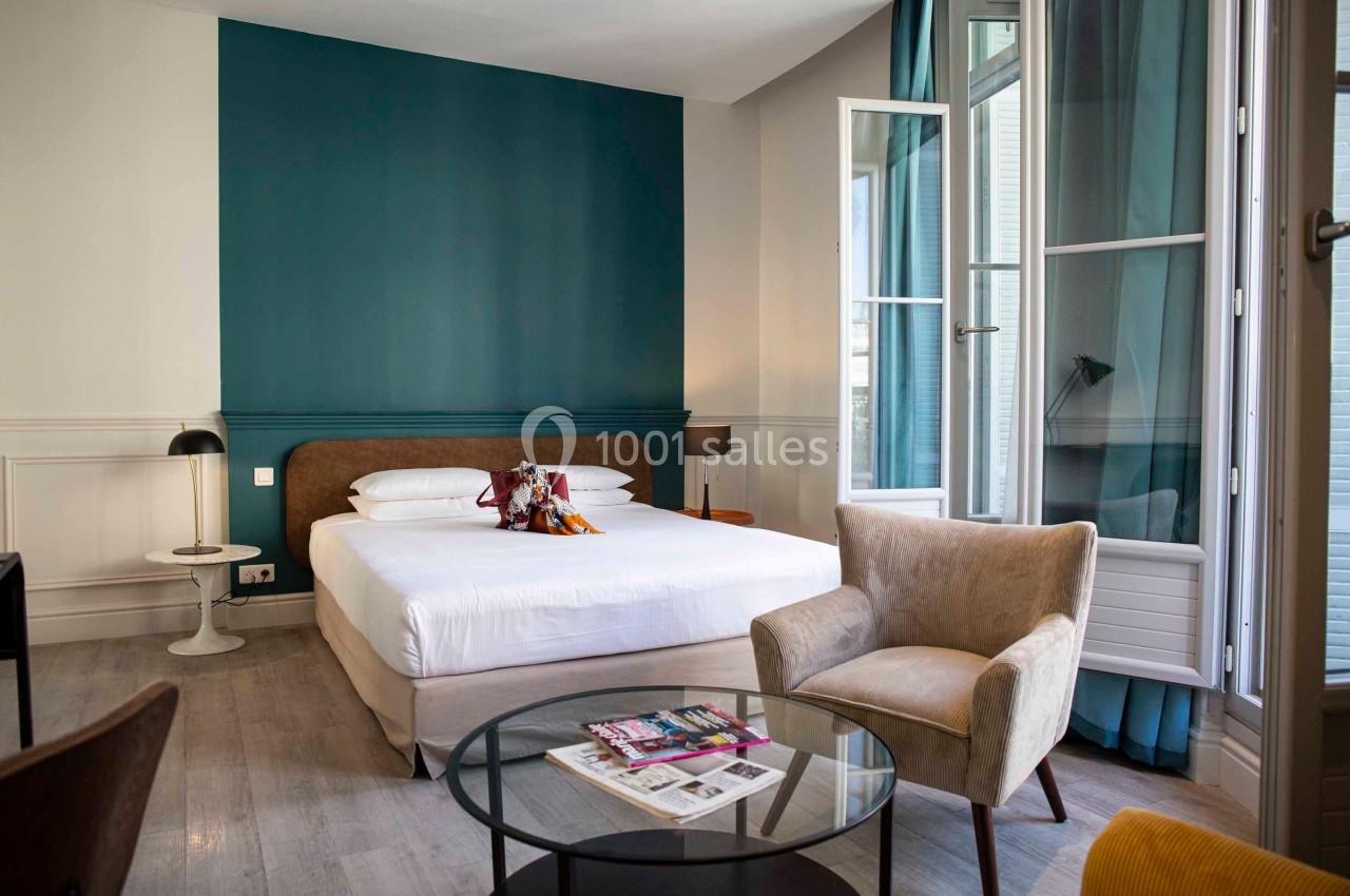 Chambre lumineuse avec lit double, fauteuil, table basse, magazines et grande fenêtre ouverte sur des rideaux bleus.