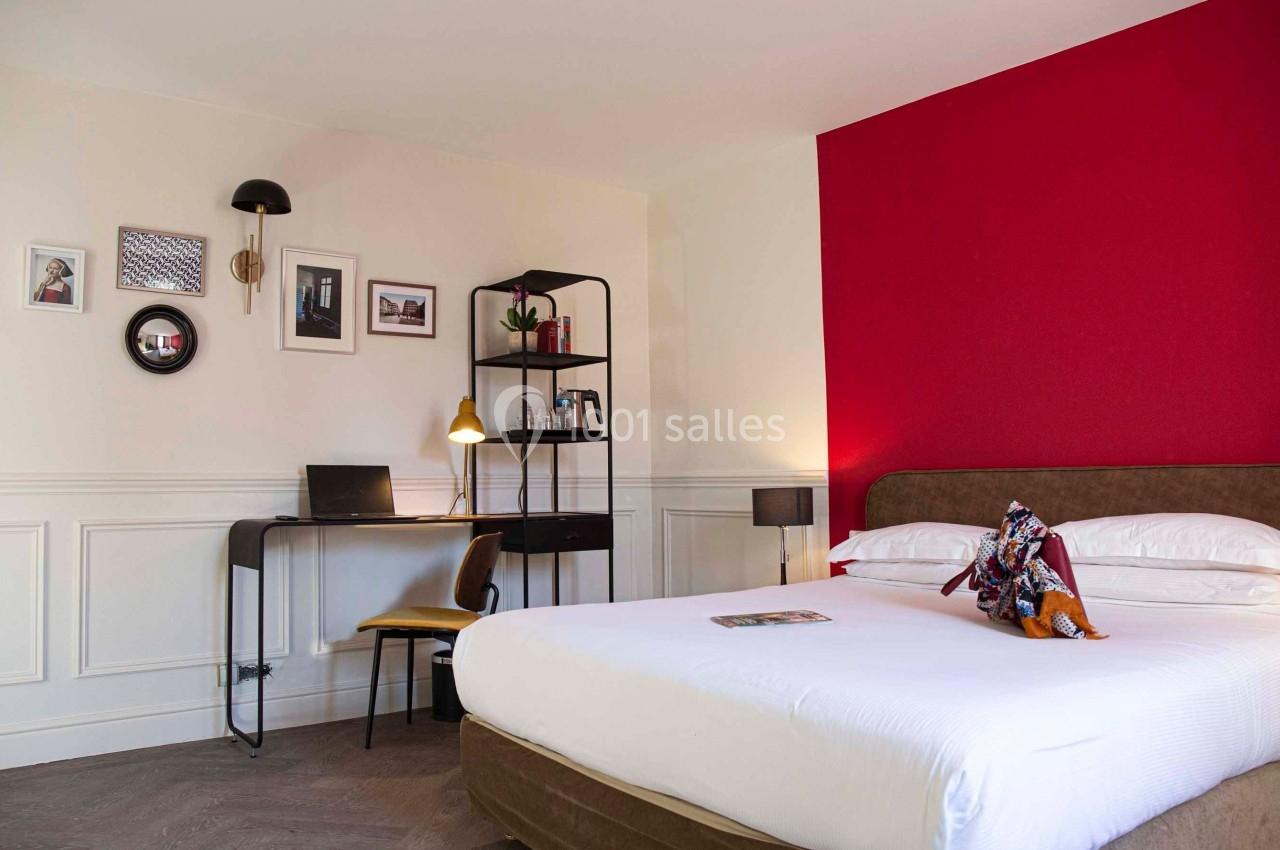 Chambre lumineuse avec un lit double, un bureau équipé d'un ordinateur portable et un mur rouge en arrière-plan.