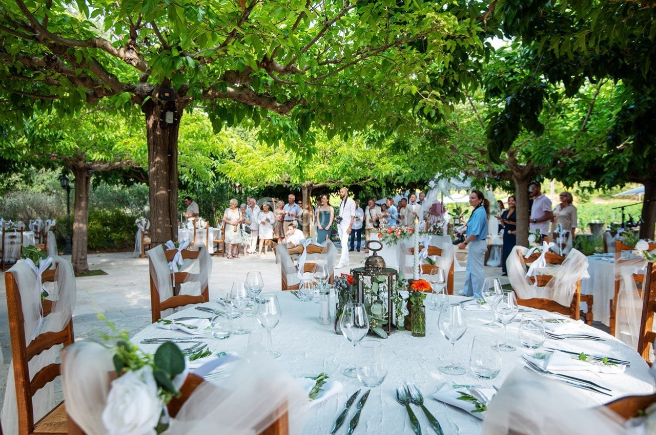 Tables décorées pour un événement en plein air sous des arbres, avec des invités rassemblés en arrière-plan.