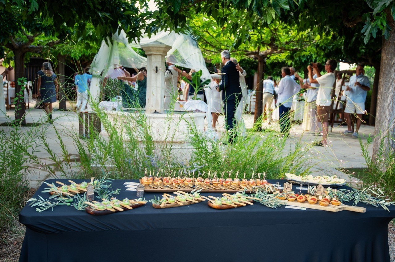 Buffet de canapés disposé sur une table noire en extérieur, avec des invités discutant en arrière-plan près d'une fontaine.