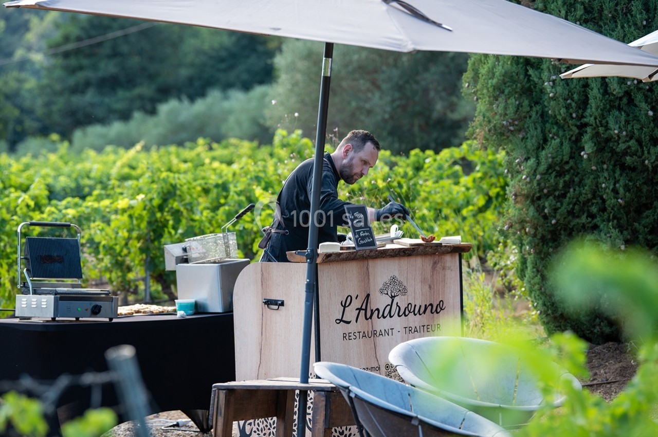 Un chef cuisine en plein air derrière un stand en bois, entouré de vignes et d'arbres sous un parasol.