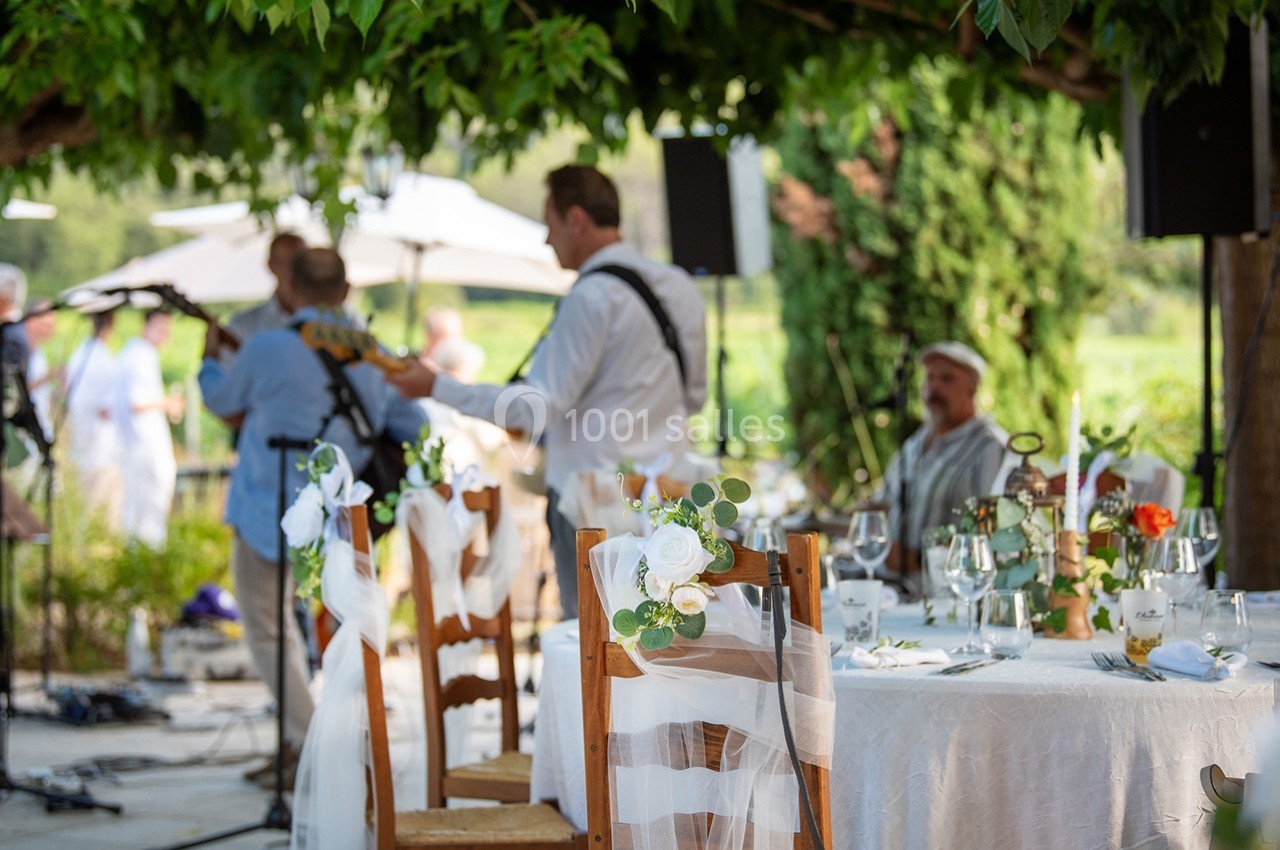 Musiciens jouant en plein air près de tables décorées pour un événement sous des arbres, avec des invités en arrière-plan.