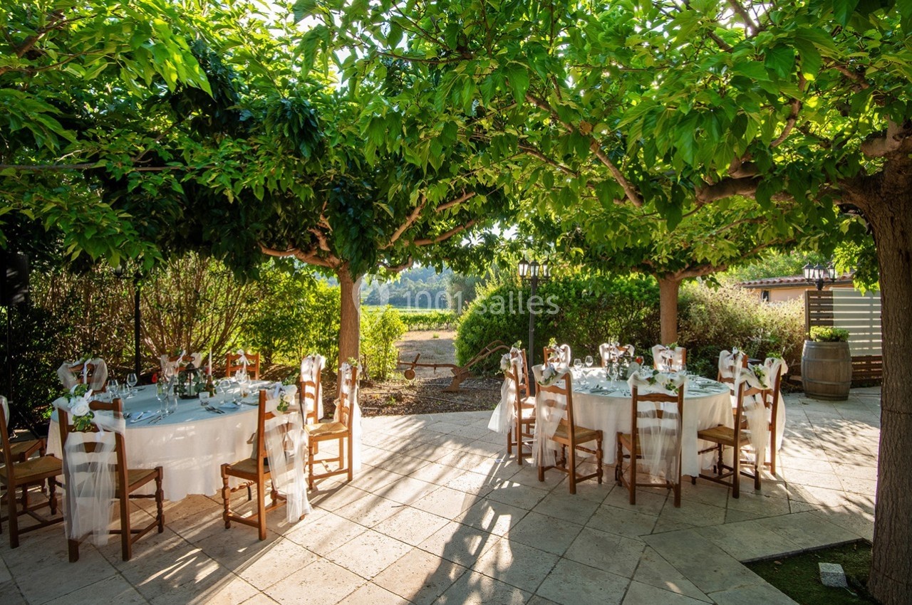Tables rondes dressées pour un repas en extérieur, disposées sous des arbres offrant une ombre naturelle.