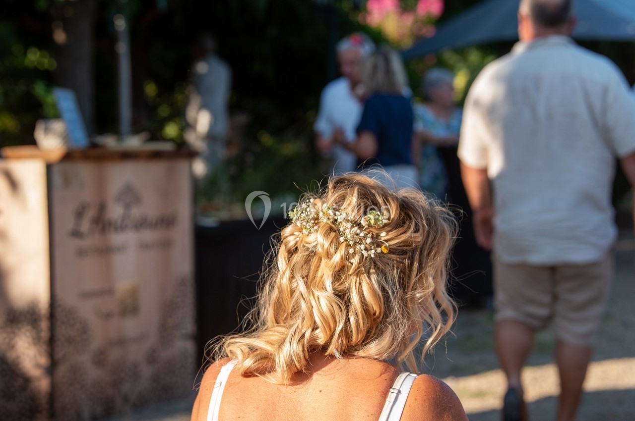 Une femme de dos avec des cheveux blonds coiffés de fleurs, dans un cadre extérieur ensoleillé avec des personnes en arrière…