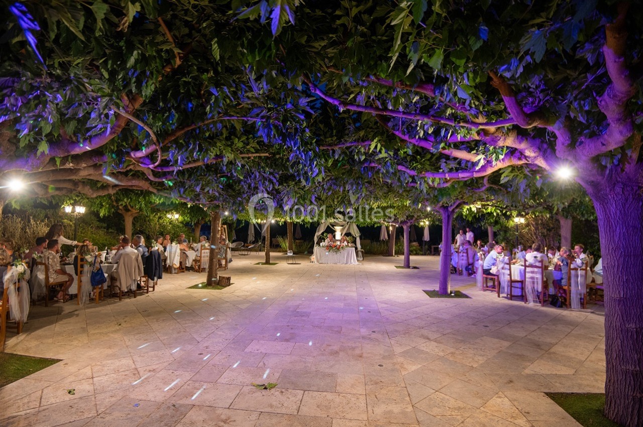 Salle de réception en plein air sous des arbres illuminés, avec des tables dressées et des invités assis.