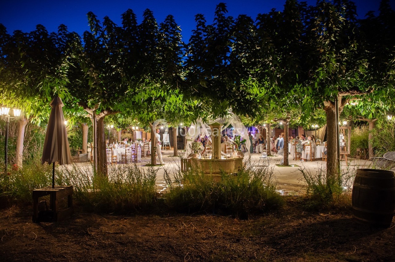 Terrasse éclairée en soirée avec des tables dressées sous des arbres, entourée de végétation et de lumières chaleureuses.