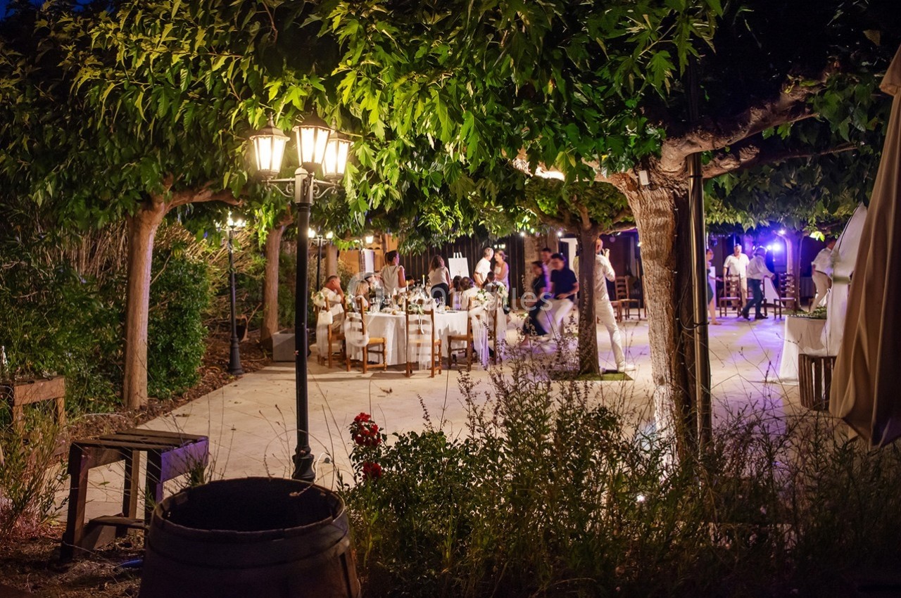 Terrasse éclairée en soirée avec des tables dressées sous des arbres, des convives assis et un éclairage chaleureux.
