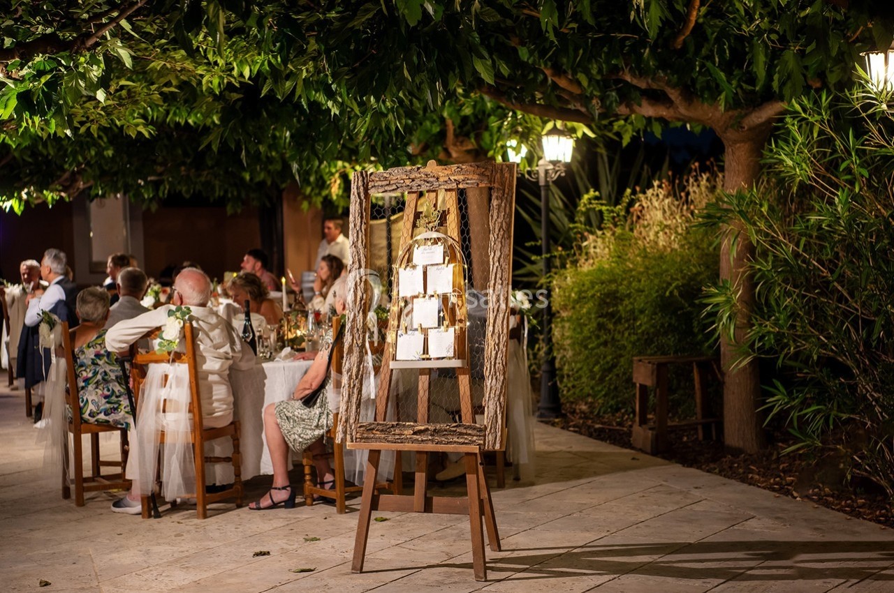 Plan de table encadré en bois devant des invités assis à des tables lors d'une réception en extérieur sous des arbres.