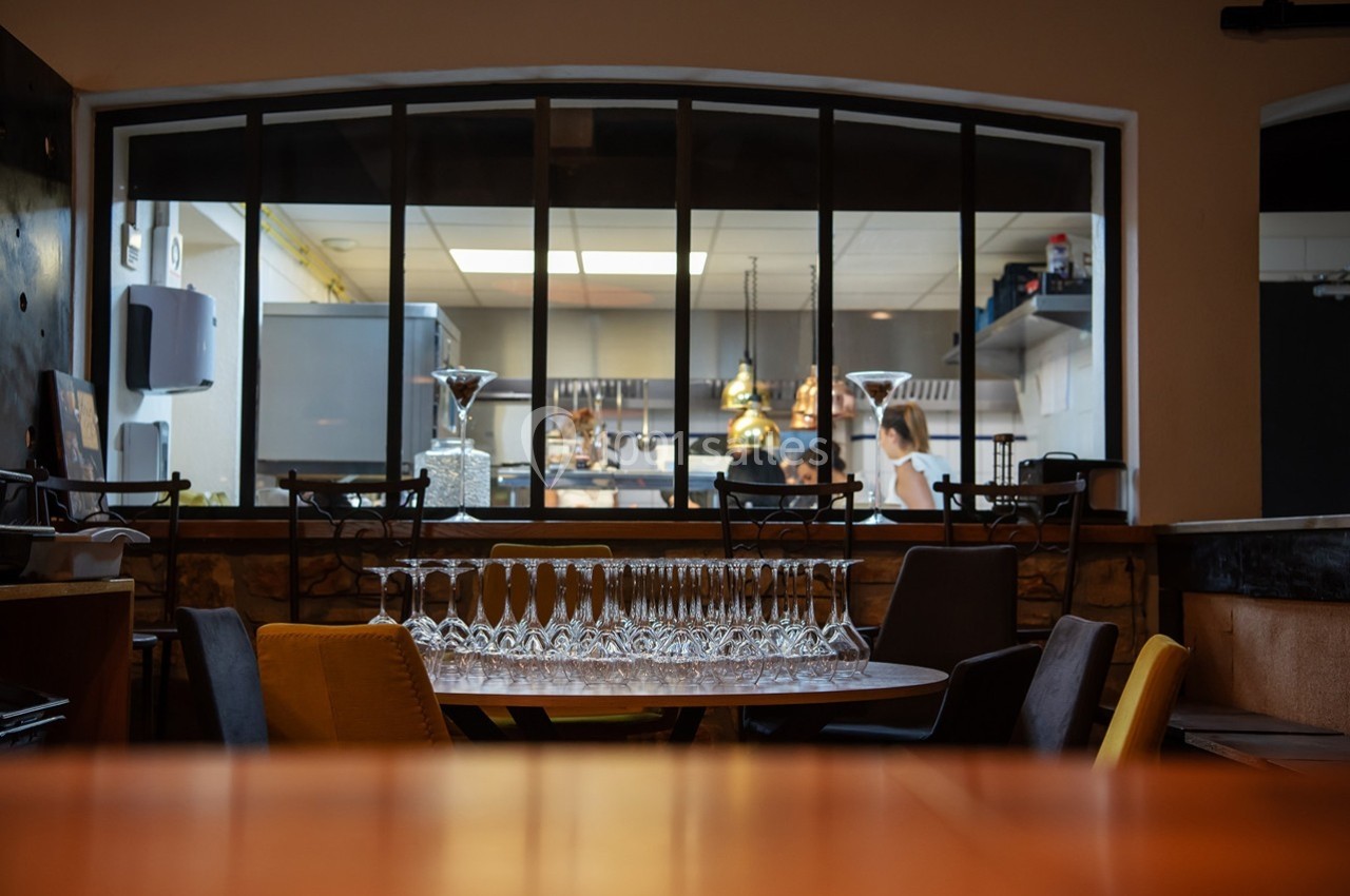 Salle de restaurant avec une table dressée de verres, donnant sur une cuisine ouverte où deux personnes travaillent.