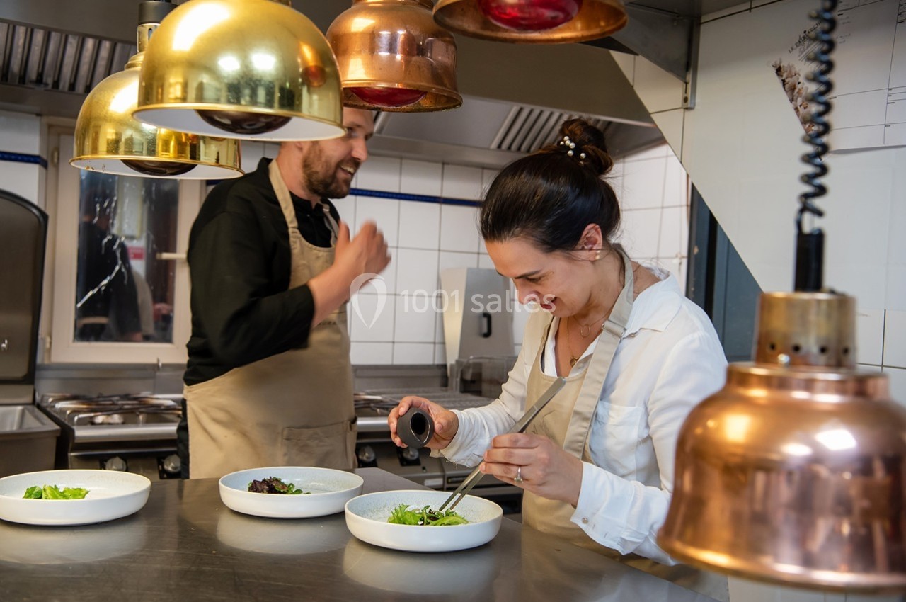Deux cuisiniers préparent des plats dans une cuisine professionnelle sous des lampes suspendues.