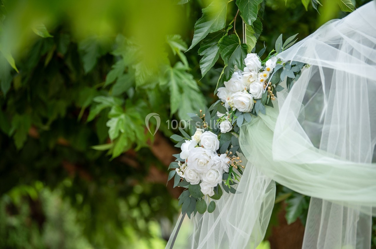 Arrangement floral blanc avec des roses et du feuillage, décoré de tulle blanc, suspendu devant un arbre verdoyant.
