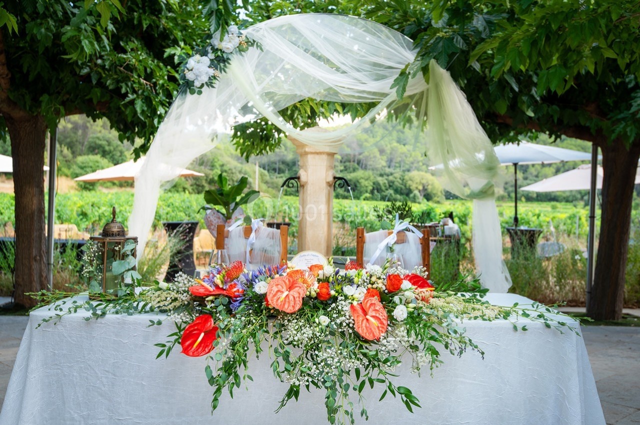 Table décorée avec des fleurs colorées et un voile blanc, installée en extérieur sous des arbres.