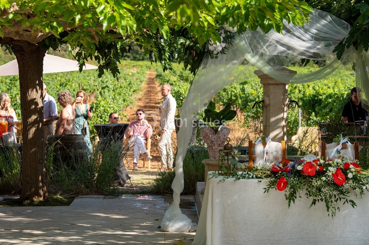 Table décorée avec des fleurs rouges et blanches sous une tonnelle, avec des invités et des vignes en arrière-plan.
