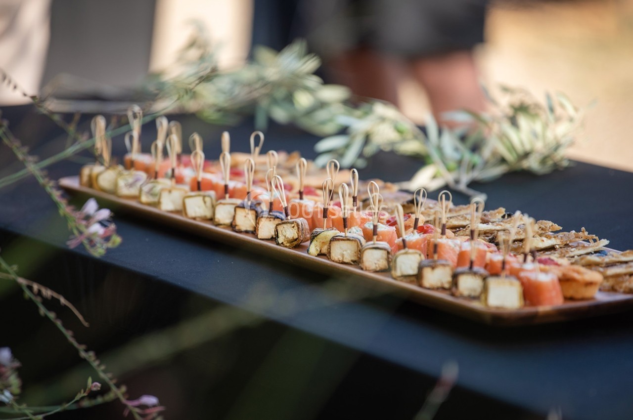 Plateau de bouchées apéritives variées disposées sur une table noire, décorée de branches d'olivier.