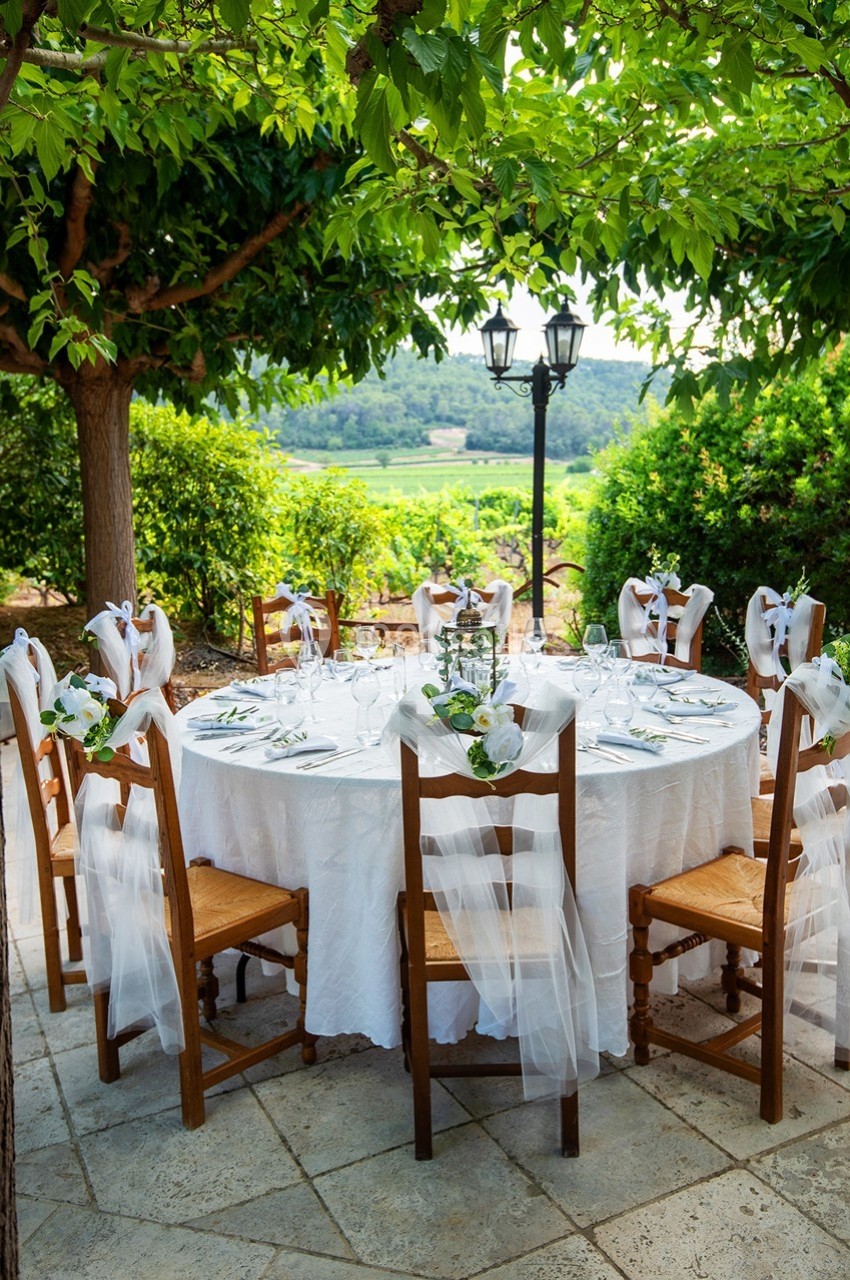 Table ronde décorée avec nappes blanches, chaises ornées de rubans et fleurs, située en extérieur sous des arbres.