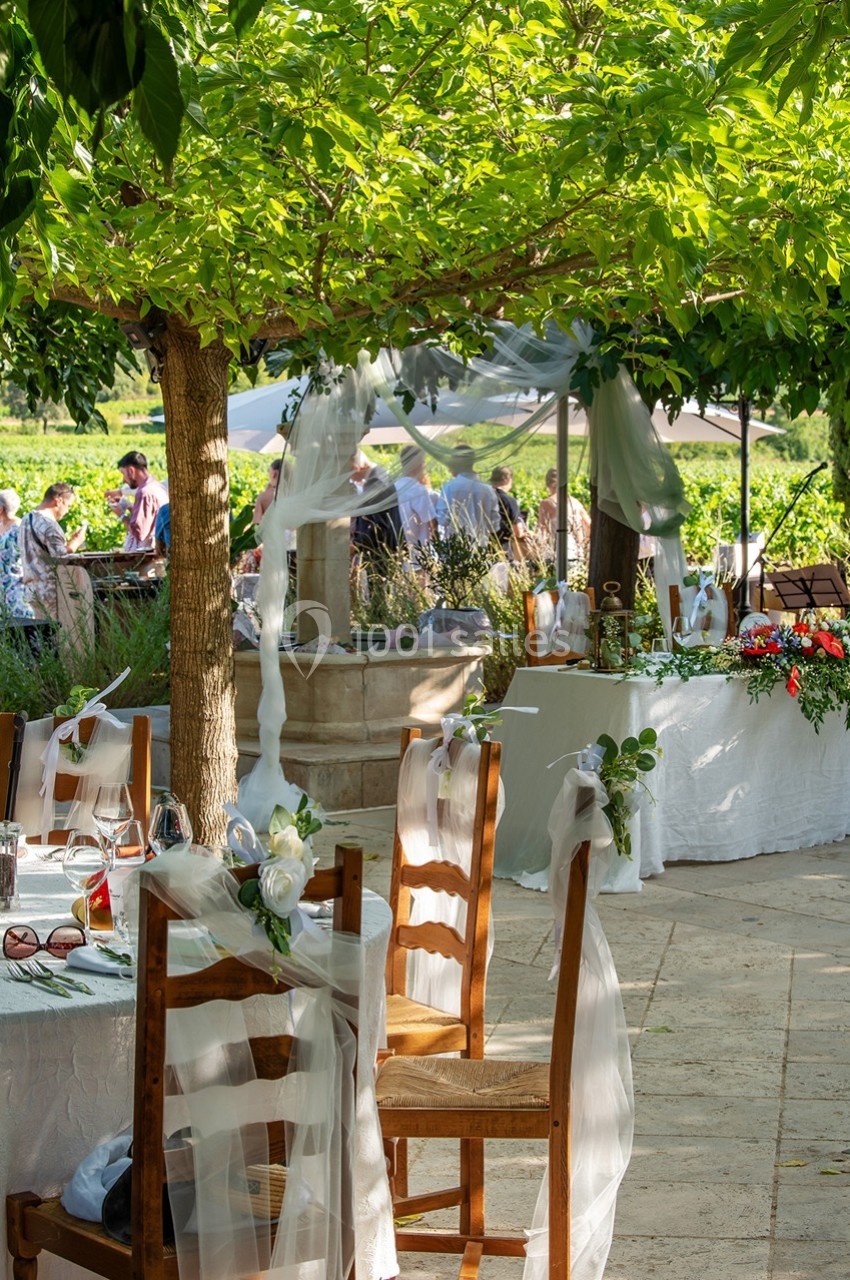 Table décorée pour un événement en plein air sous un arbre, avec nappes blanches et rubans, près d'une fontaine.