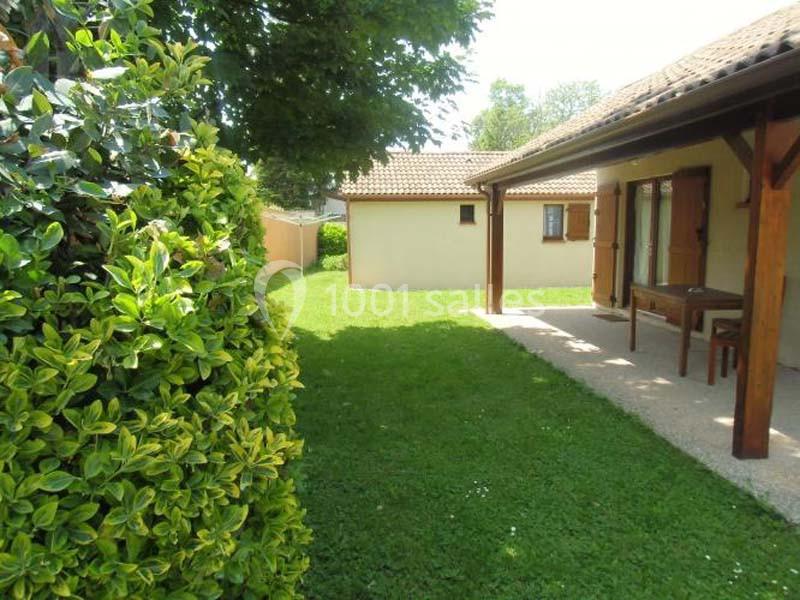 Vue d'un jardin avec pelouse, terrasse couverte, arbuste à gauche et petite dépendance au fond.