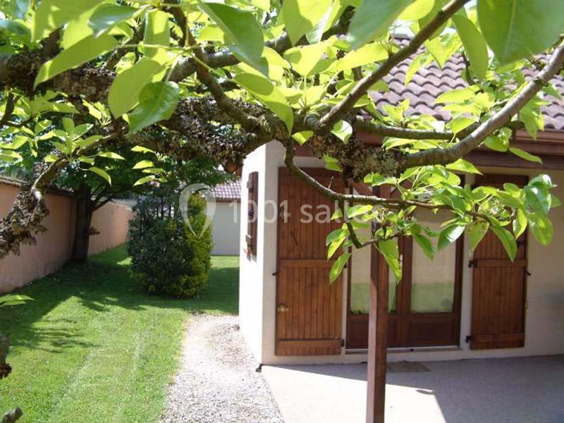 Maison avec volets en bois, entourée d'un jardin verdoyant et vue partielle d'un arbre au premier plan.