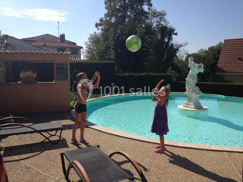Deux enfants jouent avec un ballon près d'une piscine circulaire entourée de chaises longues et d'une statue.