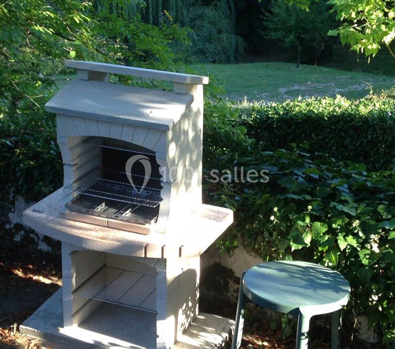 Barbecue en pierre dans un jardin verdoyant, accompagné d'un tabouret vert et entouré de haies et d'arbres.
