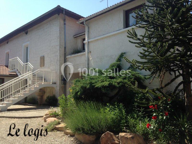 Façade d'une maison blanche avec escalier extérieur, jardin arboré et plantes fleuries au premier plan.