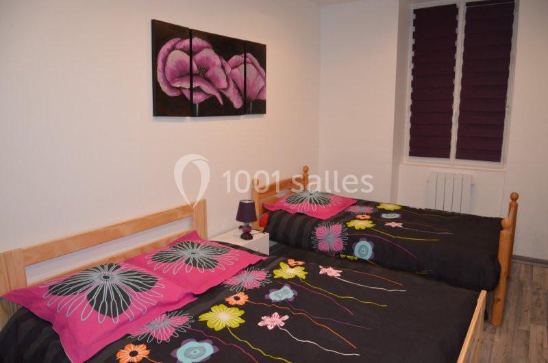 Deux lits simples avec des draps colorés, une lampe de chevet et un tableau floral au mur dans une chambre lumineuse.