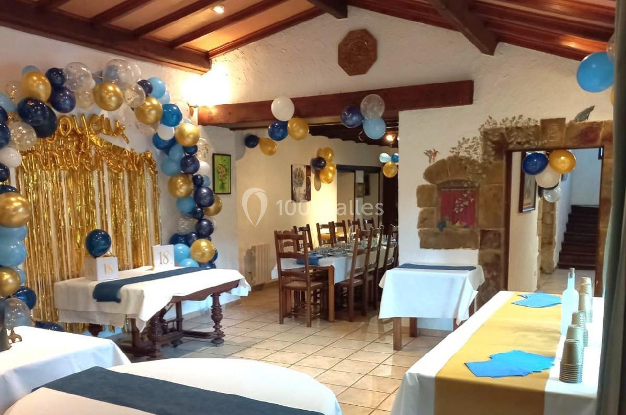 Salle décorée pour une fête, avec ballons colorés, guirlandes dorées et tables dressées pour les invités.