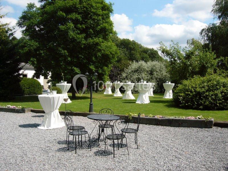 Jardin avec des tables hautes drapées de blanc et un ensemble de mobilier en fer forgé sur une allée gravillonnée.