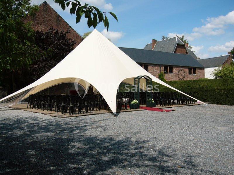 Grande tente blanche montée dans une cour pavée, entourée de bâtiments en briques et de verdure.