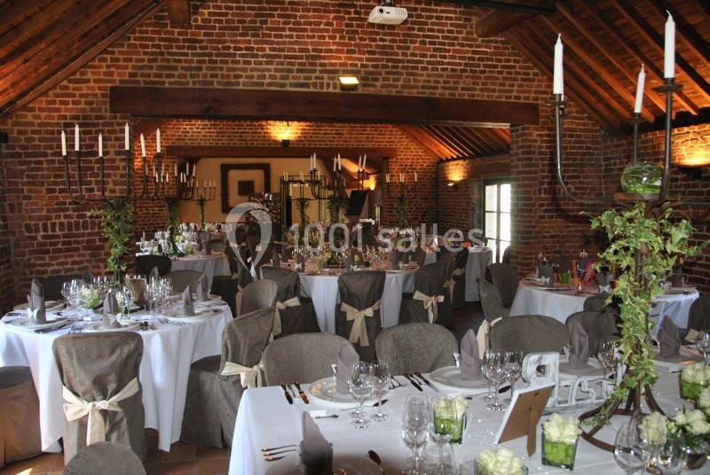 Salle de réception en briques avec tables rondes décorées, nappes blanches, chandeliers et chaises ornées de nœuds beiges.