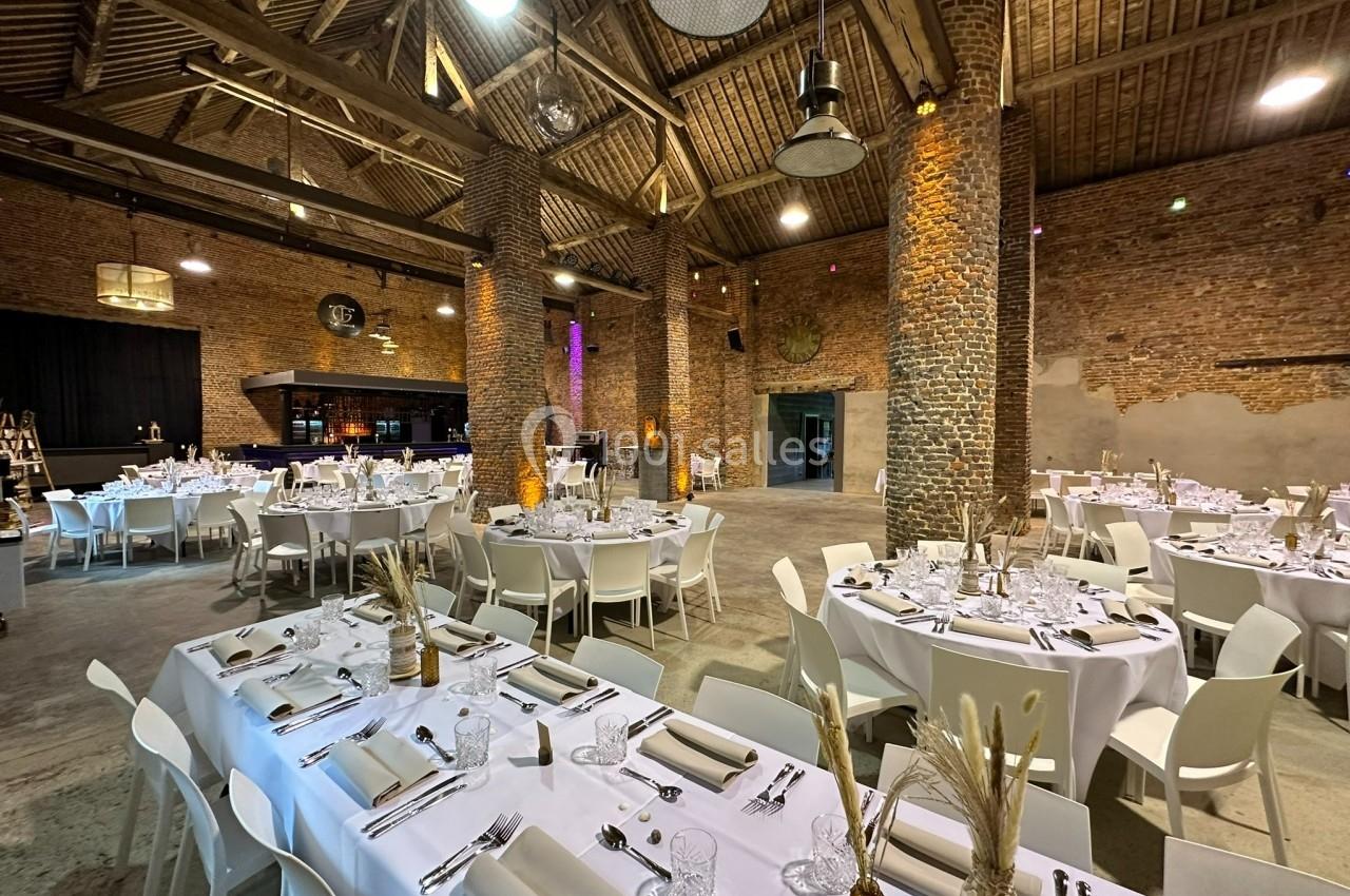 Salle de réception en briques avec tables rondes et rectangulaires dressées pour un événement.