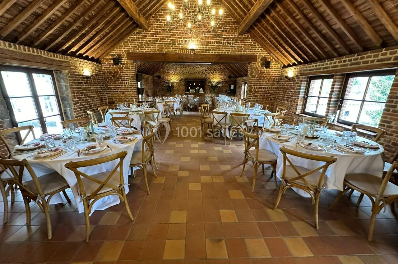 Salle de réception en briques avec tables rondes dressées, chaises en bois et lustres suspendus au plafond en poutres…