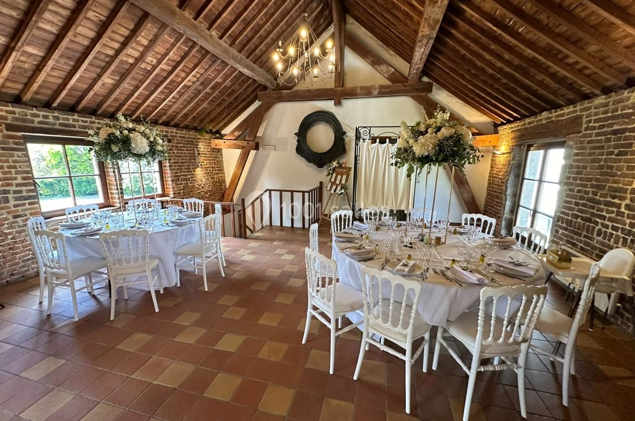 Salle rustique avec poutres apparentes, tables rondes dressées pour un repas, décorations florales et lumière naturelle.