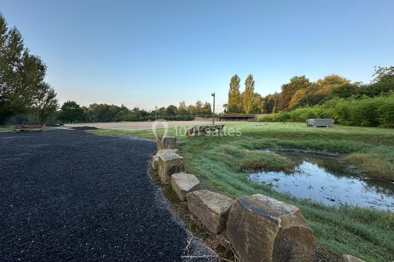 Chemin en gravier bordé de pierres, avec bancs, pelouse, étang et arbres sous un ciel dégagé en arrière-plan.