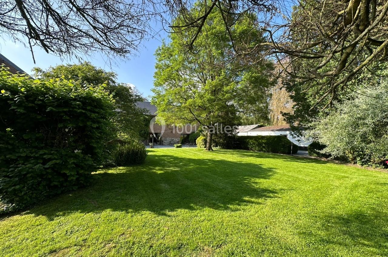 Jardin verdoyant avec pelouse bien entretenue, arbres ombragés et bâtiments en arrière-plan sous un ciel dégagé.