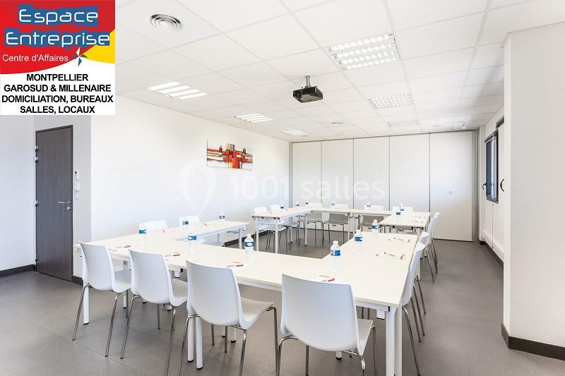 Salle de réunion lumineuse avec tables blanches disposées en U, chaises blanches et tableau mural coloré.