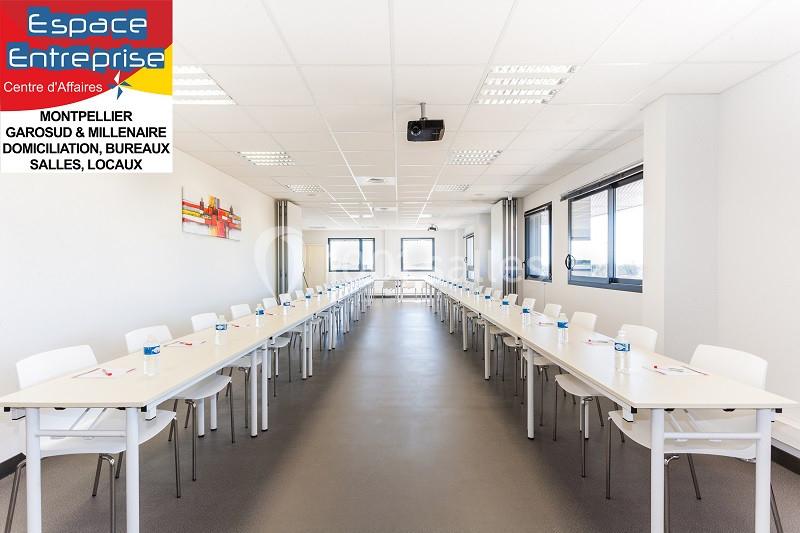 Salle de réunion lumineuse avec de longues tables alignées, chaises et fenêtres offrant une vue dégagée.